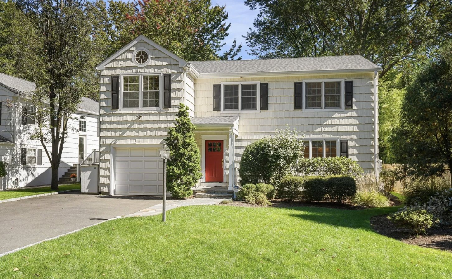 Cream shingle-sided Greenwich, Connecticut home with black shutters, a red front door under a small portico, a one-car garage, a flagstone walkway, a lawn and mature trees