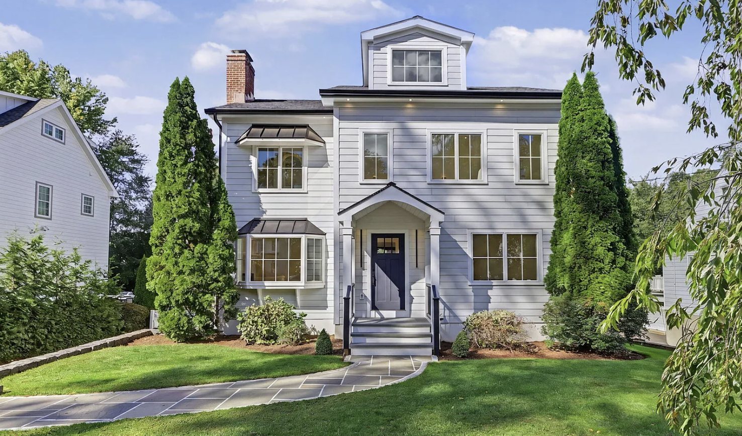 White-sided Greenwich, Connecticut home with a dormer, bay windows, a navy front door under a gabled portico, a flagstone walkway and tall evergreens