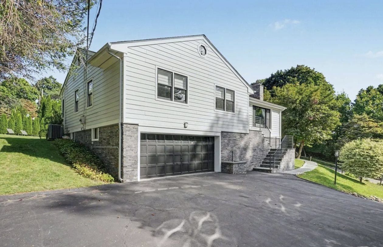 Gray-sided Greenwich, Connecticut home with a dark two-car garage, stone base, and a stairway with black railings up to the entrance